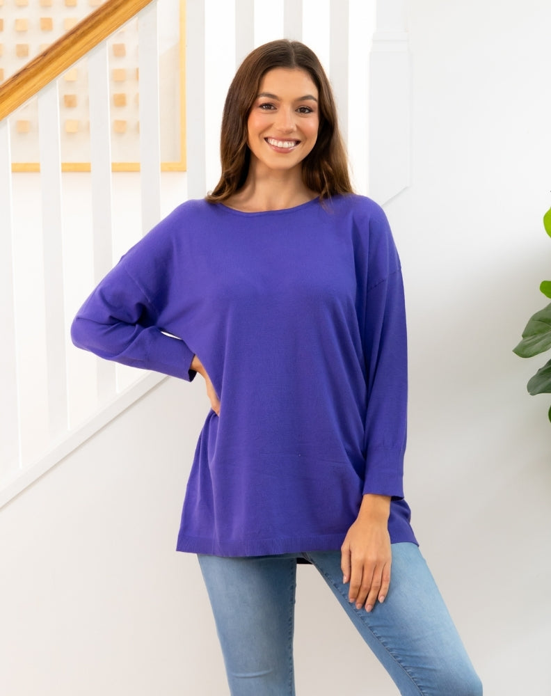 Woman wearing a purple long-sleeve top and blue jeans standing indoors.