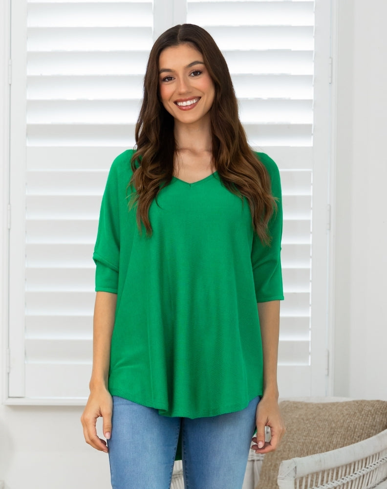 A woman wearing a bright green three-quarter sleeve V-neck top and blue jeans, standing in front of white window shutters.