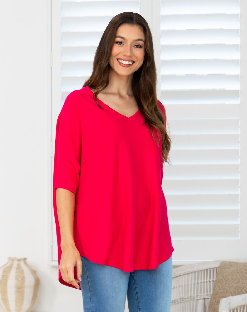 A woman wearing a bright red V-neck top with three-quarter sleeves and blue jeans, standing in front of white window shutters.