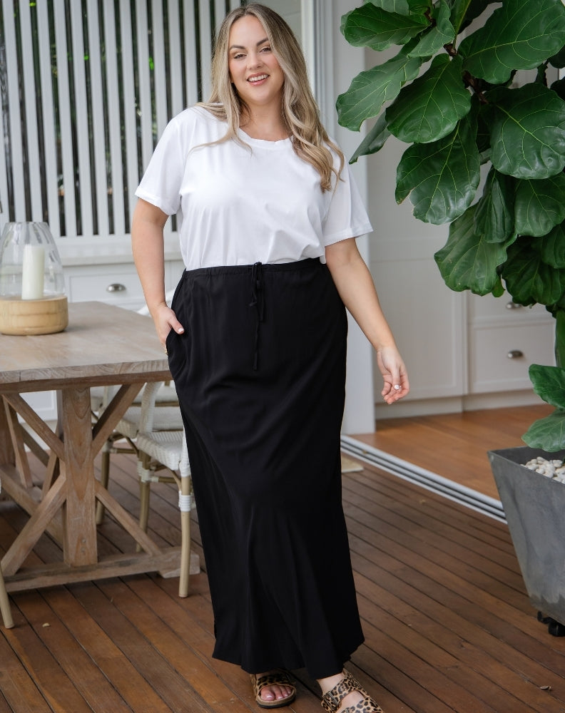 Woman wearing a white top and black skirt standing on a wooden deck with plants and furniture in the background.