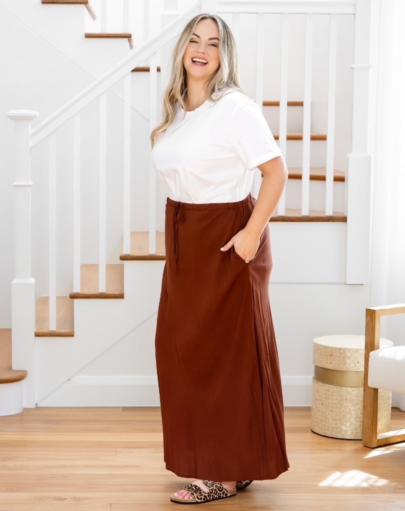 Woman wearing a white top and brown skirt standing in a home setting with stairs in the background.