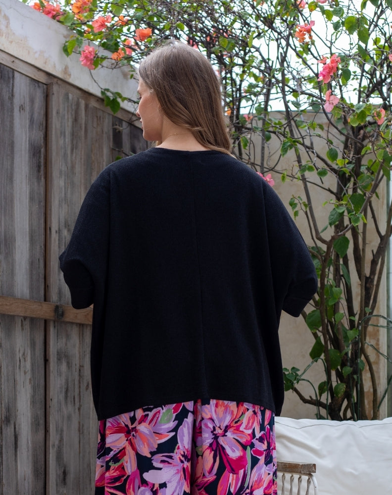 Person wearing a black top and colorful floral skirt standing outdoors with plants and flowers in the background.