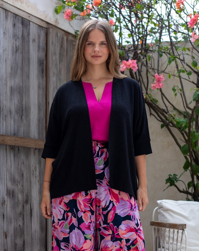 Woman wearing a black cardigan over a pink top and floral pants in an outdoor setting.