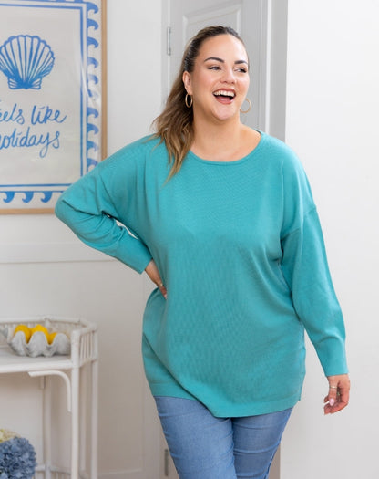 Woman wearing a teal sweater and blue jeans standing in a room with a decorative wall sign.