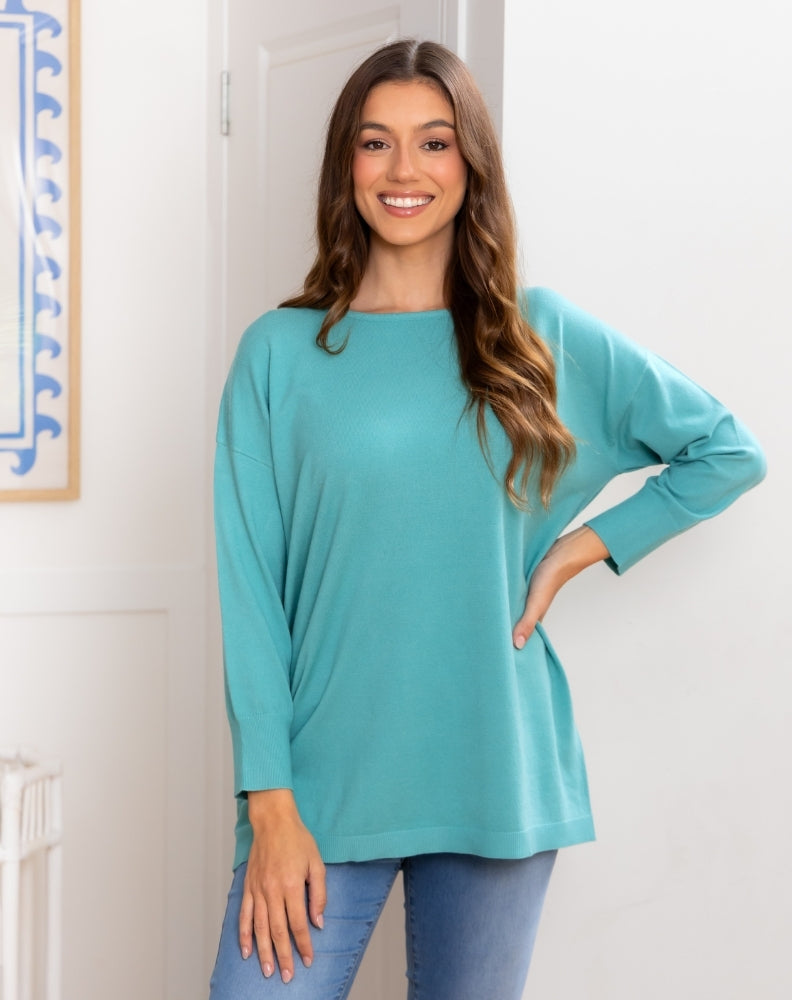 Woman wearing a teal long-sleeve top and blue jeans indoors.