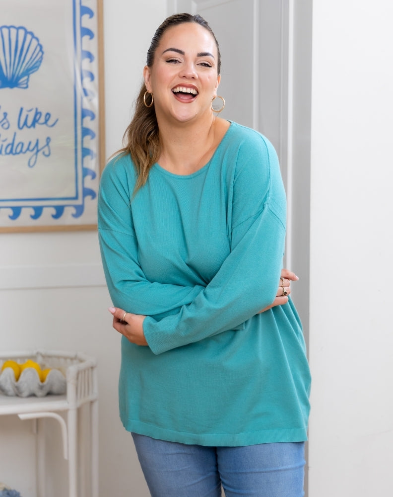 Woman wearing a teal sweater and blue jeans standing in a room with a decorative wall sign.