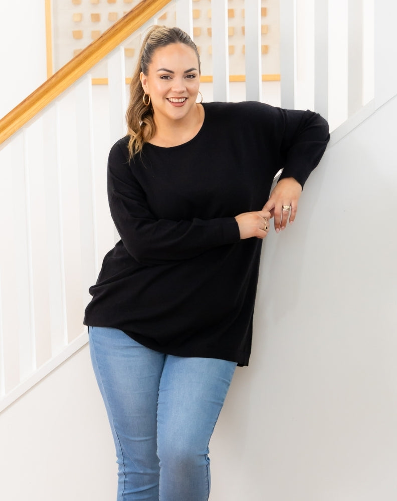 Woman wearing a black oversized sweater and light blue jeans standing against a white wall.