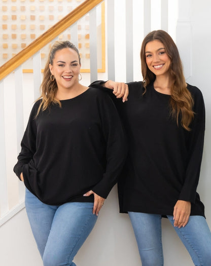 Two women wearing black oversized tunic knit and blue jeans standing against a white wall.