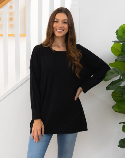 Woman wearing a black long-sleeve top and blue jeans standing indoors with a plant in the background.