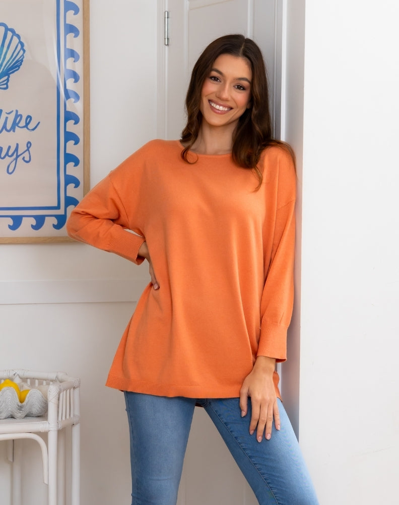 Woman wearing an orange long-sleeve top and blue jeans standing in a room with a decorative wall sign.