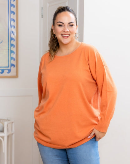 Woman wearing an orange long-sleeve top and blue jeans indoors.