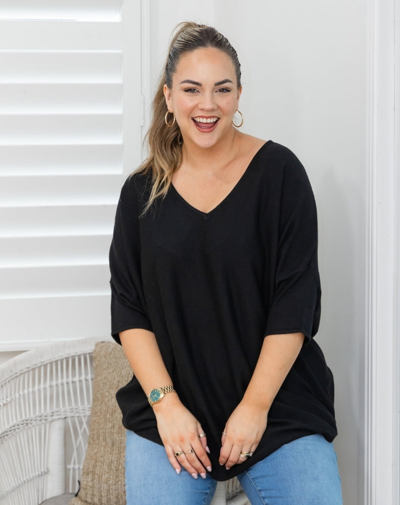 A woman wearing a black V-neck, three-quarter sleeve top and light blue jeans, smiling in front of a white wall with window blinds.