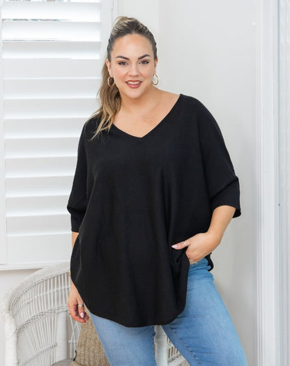 A woman wearing a black V-neck, three-quarter sleeve top and light blue jeans, standing in front of white window shutters.