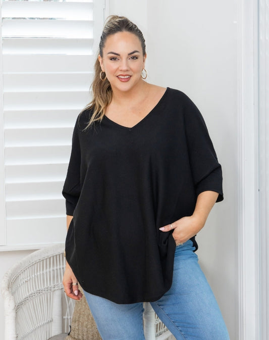 A woman wearing a black V-neck, three-quarter sleeve top and light blue jeans, standing in front of white window shutters.