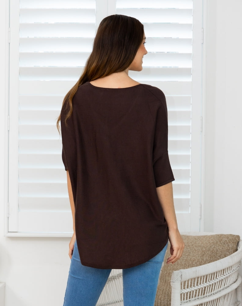 A woman shown from the back wearing a dark brown, loose-fitting short-sleeve top and blue jeans, standing in front of white window shutters.