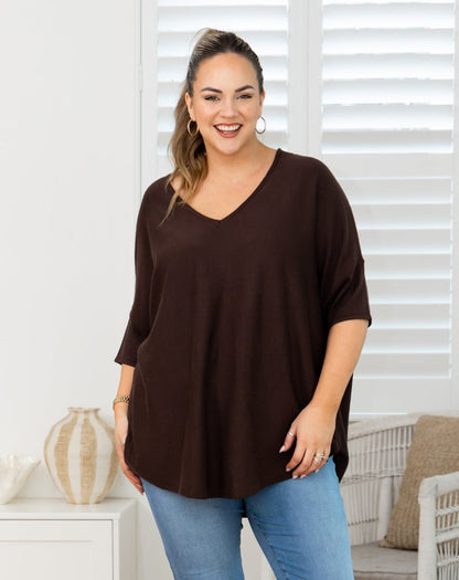 A woman modeling a dark brown V-neck short-sleeve top with blue jeans in a bright, modern room setting.