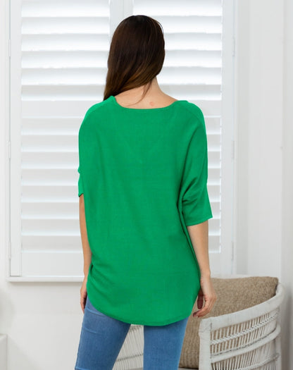 A woman viewed from the back wearing a bright green, loose-fitting short-sleeve top and blue jeans, standing in front of white window shutters.