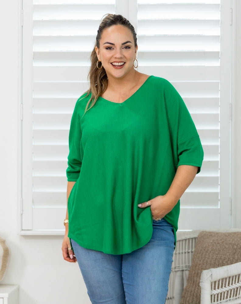 A woman wearing a bright green, loose-fitting three-quarter sleeve top and blue jeans, standing in front of white window shutters.