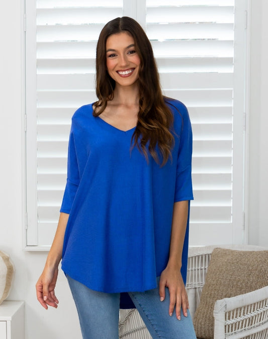 A woman wearing a bright blue V-neck tunic top with three-quarter sleeves and light-wash blue jeans, standing in front of white window shutters.