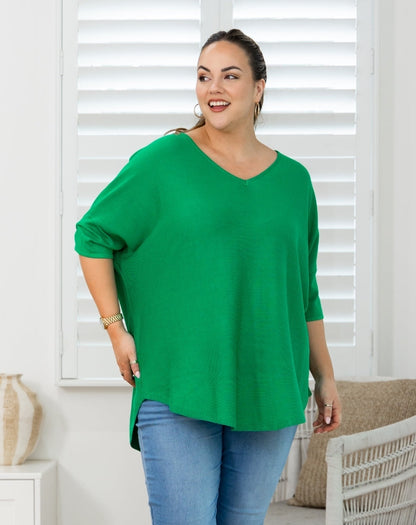 A woman modeling a bright green V-neck top with three-quarter sleeves and a side slit, paired with blue jeans, standing in front of white window shutters.