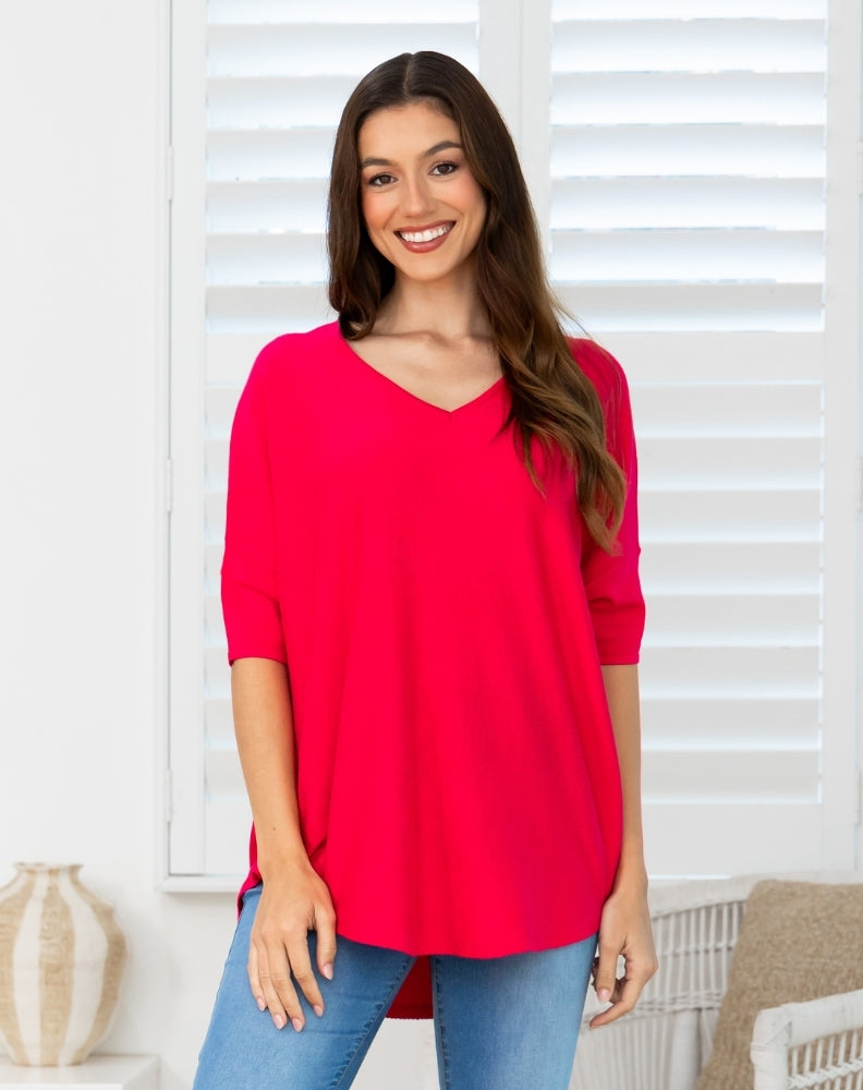 A woman wearing a bright red V-neck tunic top with three-quarter sleeves and blue jeans, standing in front of white window shutters.