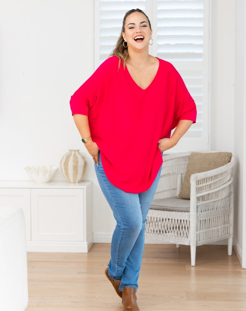 A woman modeling a bright red, oversized V-neck top with three-quarter sleeves, paired with blue jeans and brown boots, standing in a bright, modern room with white shutters and a wicker chair.