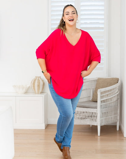 A woman modeling a bright red, oversized V-neck top with three-quarter sleeves, paired with blue jeans and brown boots, standing in a bright, modern room with white shutters and a wicker chair.