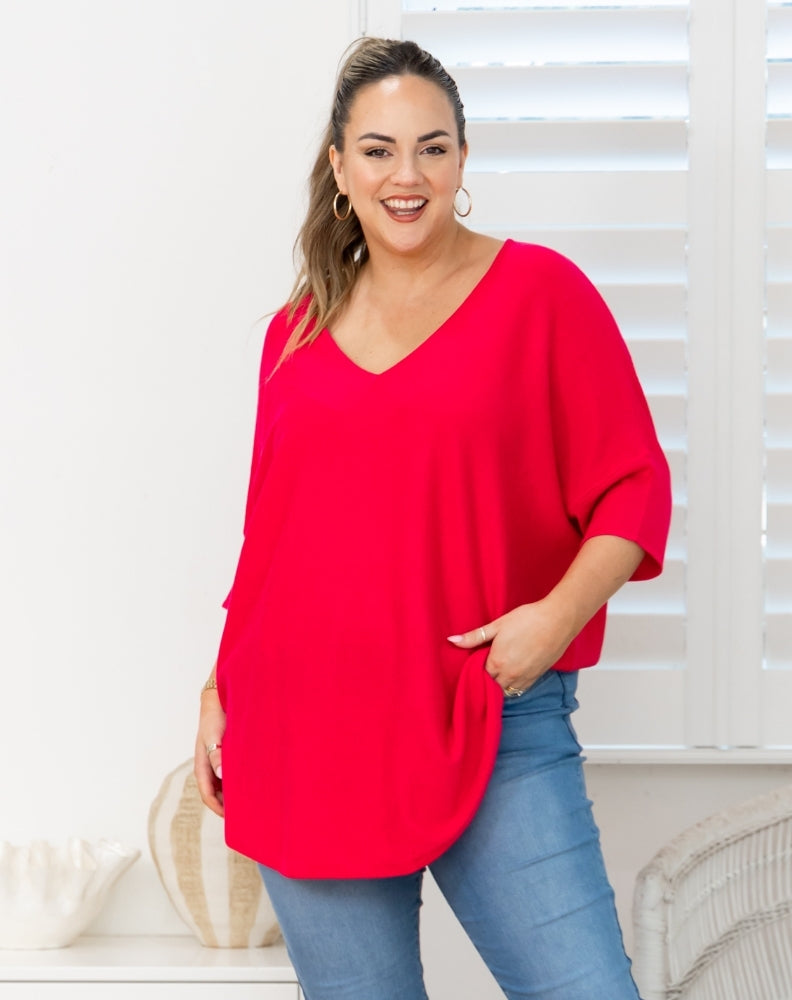 A woman wearing a bright red, oversized V-neck top with elbow-length sleeves and side pockets, paired with blue jeans, standing in front of white window blinds.