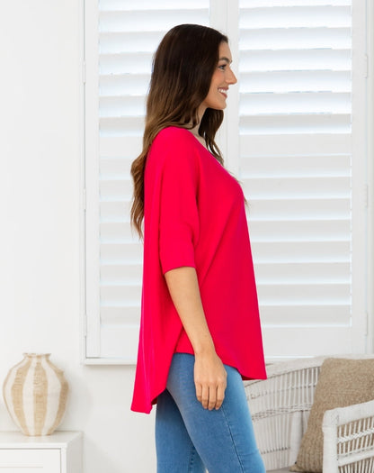 A woman wearing a bright pinkish-red short-sleeve V-neck top and blue jeans, standing in profile in front of white window shutters.