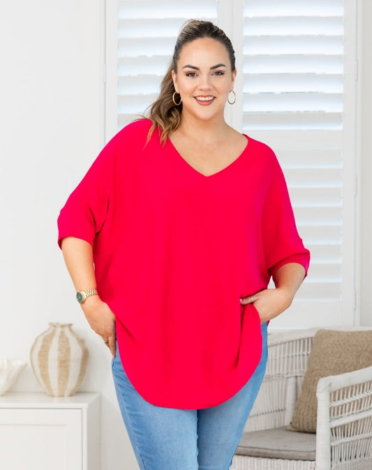 A woman wearing a bright red V-neck top with three-quarter sleeves and side pockets, paired with blue jeans, standing in a brightly lit room with white shutters and a wicker chair.