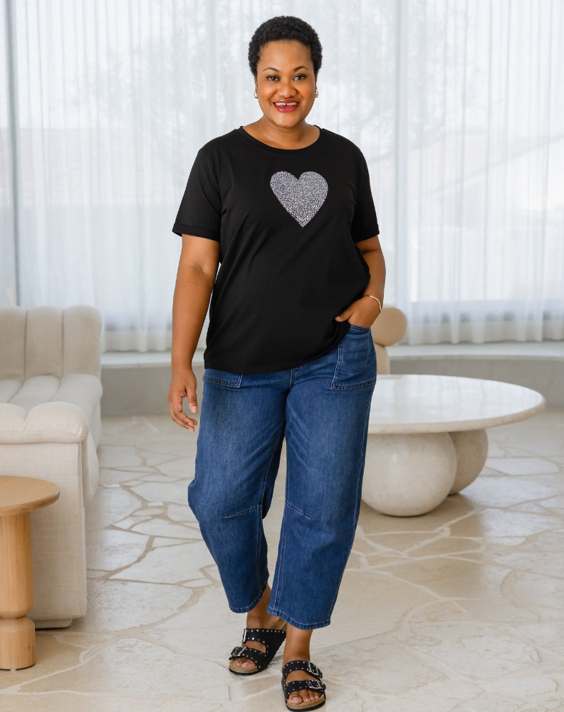 Woman wearing a black t-shirt with a heart design and blue jeans standing in a modern living room.