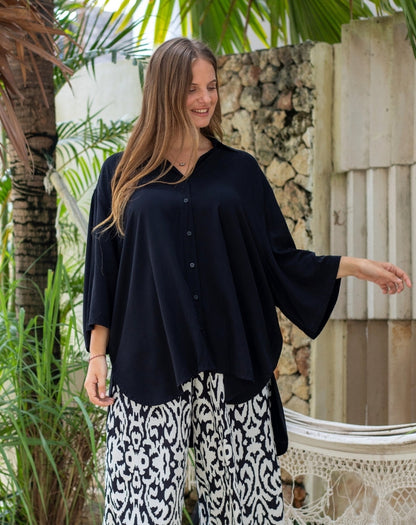 Woman wearing a black top and patterned pants standing outdoors with greenery in the background