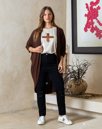 Woman standing indoors wearing a brown cardigan, white t-shirt with a cross design, black pants, and white sneakers. Behind her is a wall with a piece of abstract art.