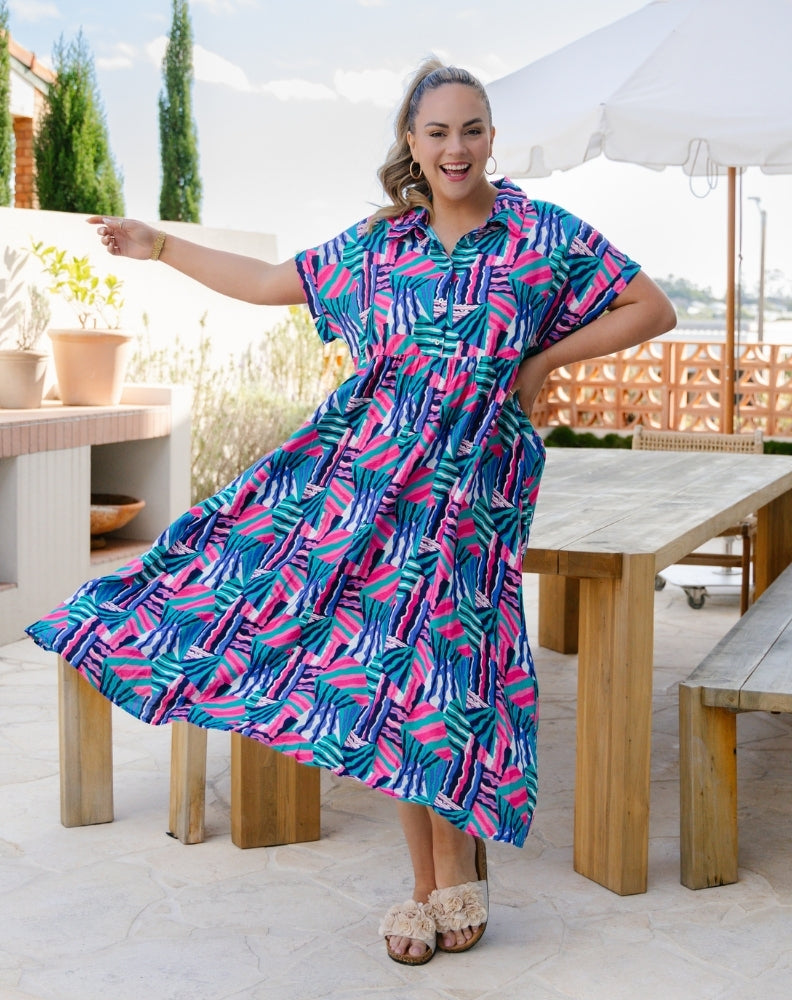 A woman modeling a short-sleeve button-up maxi dress with a colorful palm leaf pattern in pink, teal, and blue, set against an outdoor patio background with wooden tables and a white umbrella.