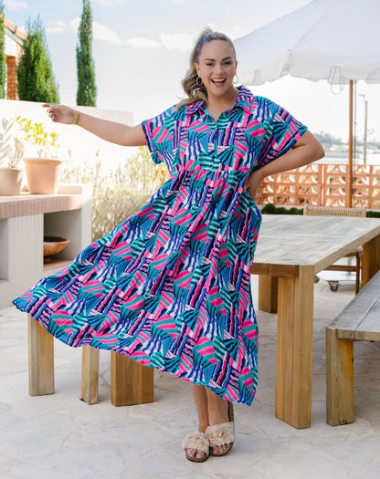 A woman modeling a short-sleeve button-up maxi dress with a colorful palm leaf pattern in pink, teal, and blue, set against an outdoor patio background with wooden tables and a white umbrella.