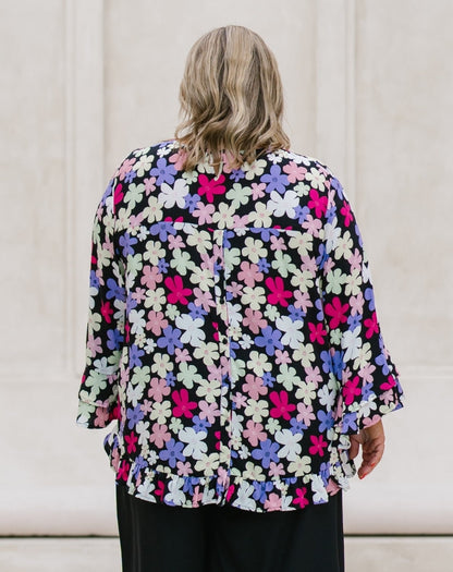 A woman viewed from behind wearing a black blouse with a vibrant multicolor floral pattern in pink, purple, and white, paired with black trousers, set against a neutral cream-colored wall.