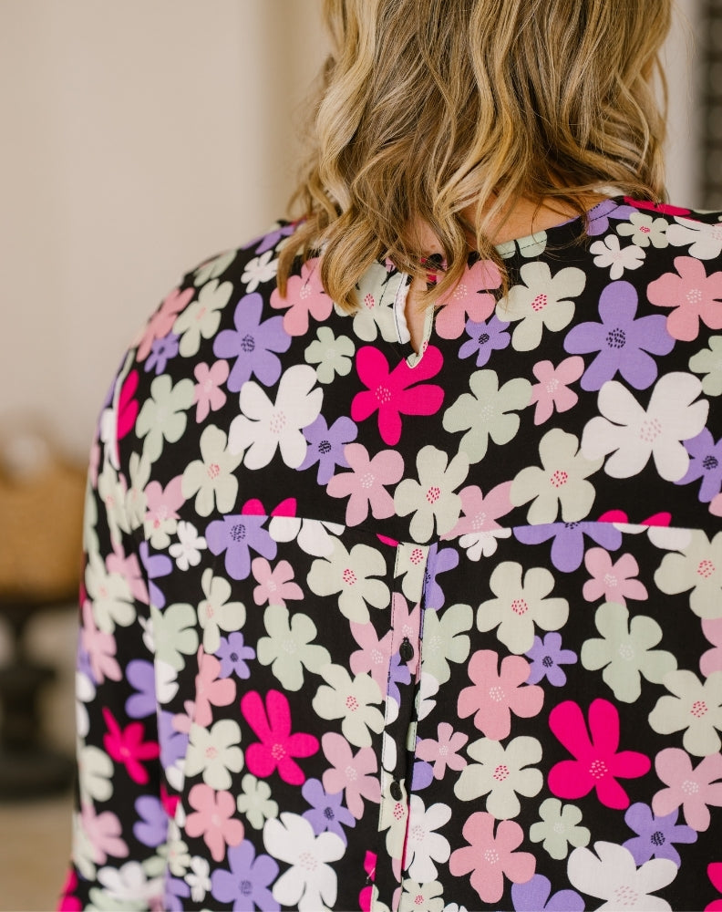 A close-up back view of a woman wearing a black long-sleeve blouse with a colorful floral pattern of pink, purple, white, and light green flowers.