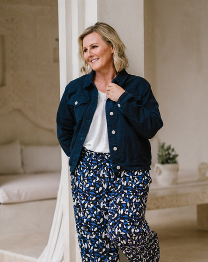 A woman wearing a dark navy blue denim jacket over a white top and blue-and-white patterned pants, standing in a softly lit indoor setting with neutral walls and furniture.