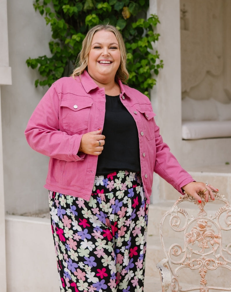 A woman smiling while wearing a bright pink cropped denim jacket over a black top and black pants with a colorful floral print, standing outdoors next to a white ornate chair.
