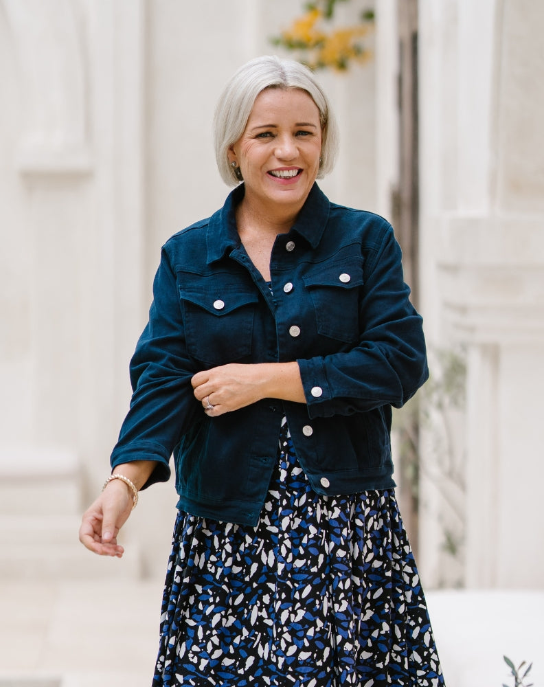 A woman with short gray hair smiling outdoors, wearing a dark teal corduroy jacket over a navy, white, and blue patterned midi skirt.