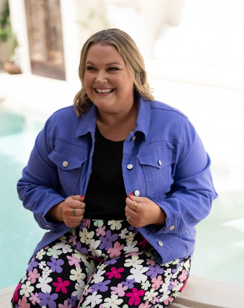 A woman smiling while wearing a blue denim-style jacket over a black top and colorful floral pants, posed outdoors near a light-colored poolside setting.