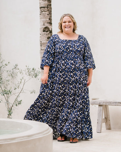 A woman modeling a navy blue maxi dress with a white abstract floral pattern, standing in a bright, modern outdoor setting with a white wall, a bench, and a birch tree trunk.
