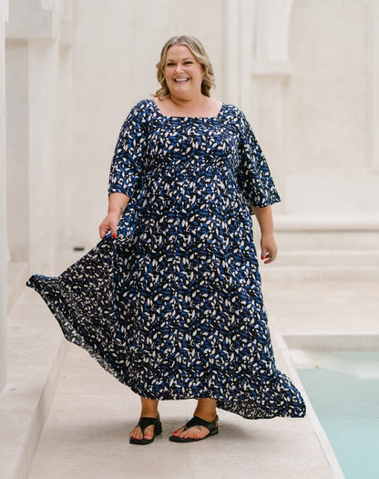 A woman wearing a navy blue and white floral print maxi dress with elbow-length sleeves, walking outdoors next to a light-colored stone structure and a pool.