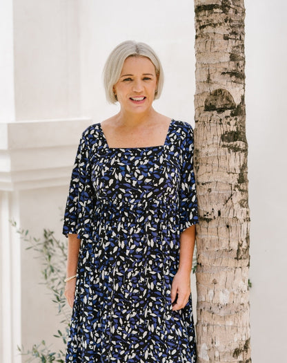 A woman with short gray hair wearing a navy blue dress with a white and light blue abstract pattern, standing next to a tree trunk in front of a white building.
