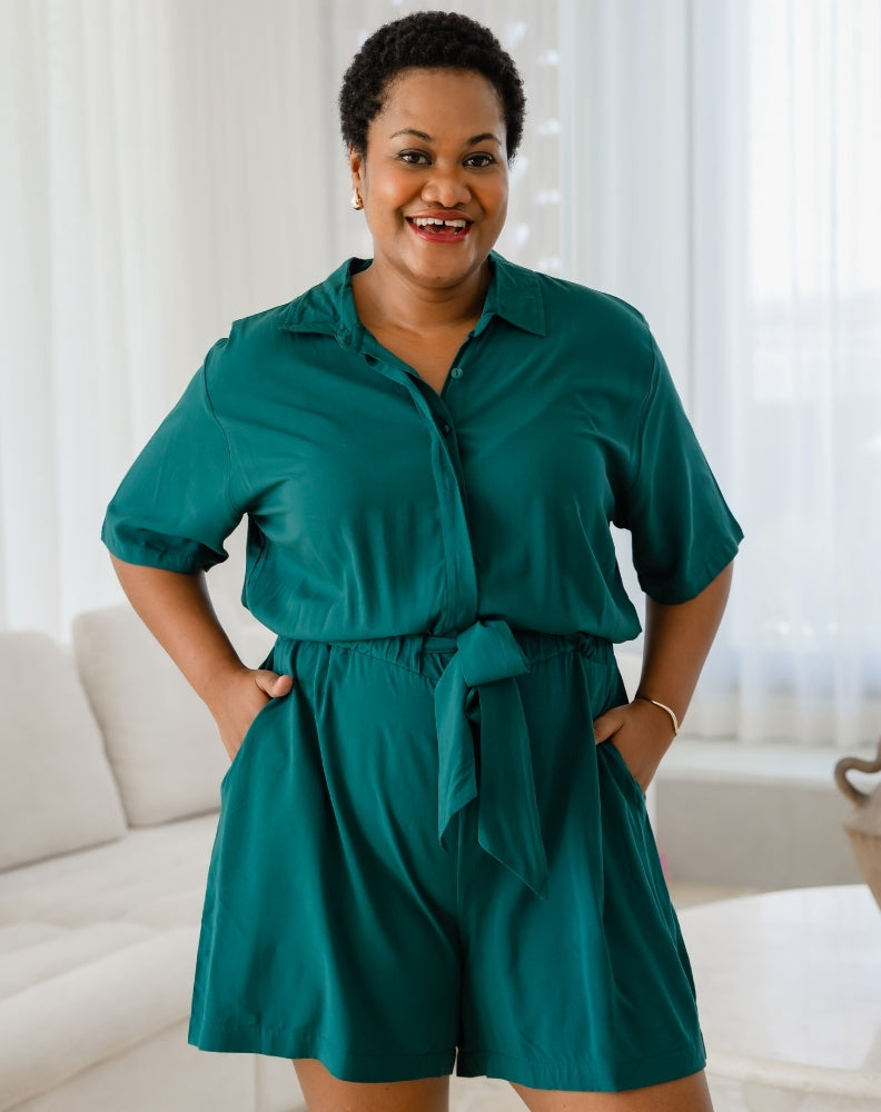 Woman wearing a teal romper standing in a bright room with white curtains.
