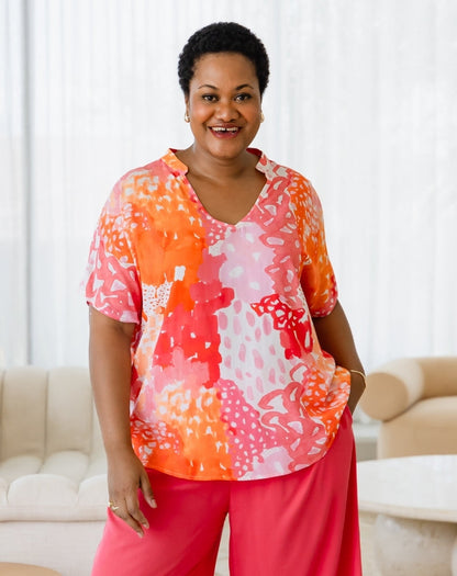 Woman wearing a colorful patterned top and pink pants in a living room setting.