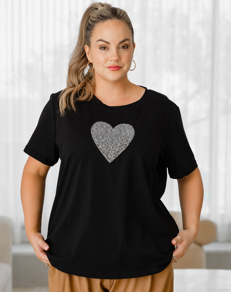 Woman wearing a black t-shirt with a silver heart design in a bright room.