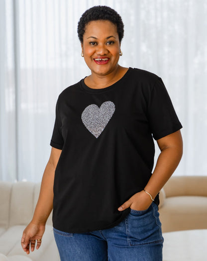 Woman wearing a black t-shirt with a heart design, standing in a room with white curtains.