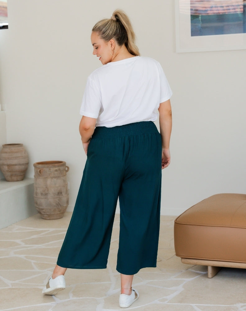 Person wearing a white t-shirt and teal pants in a room with beige walls and a brown ottoman.