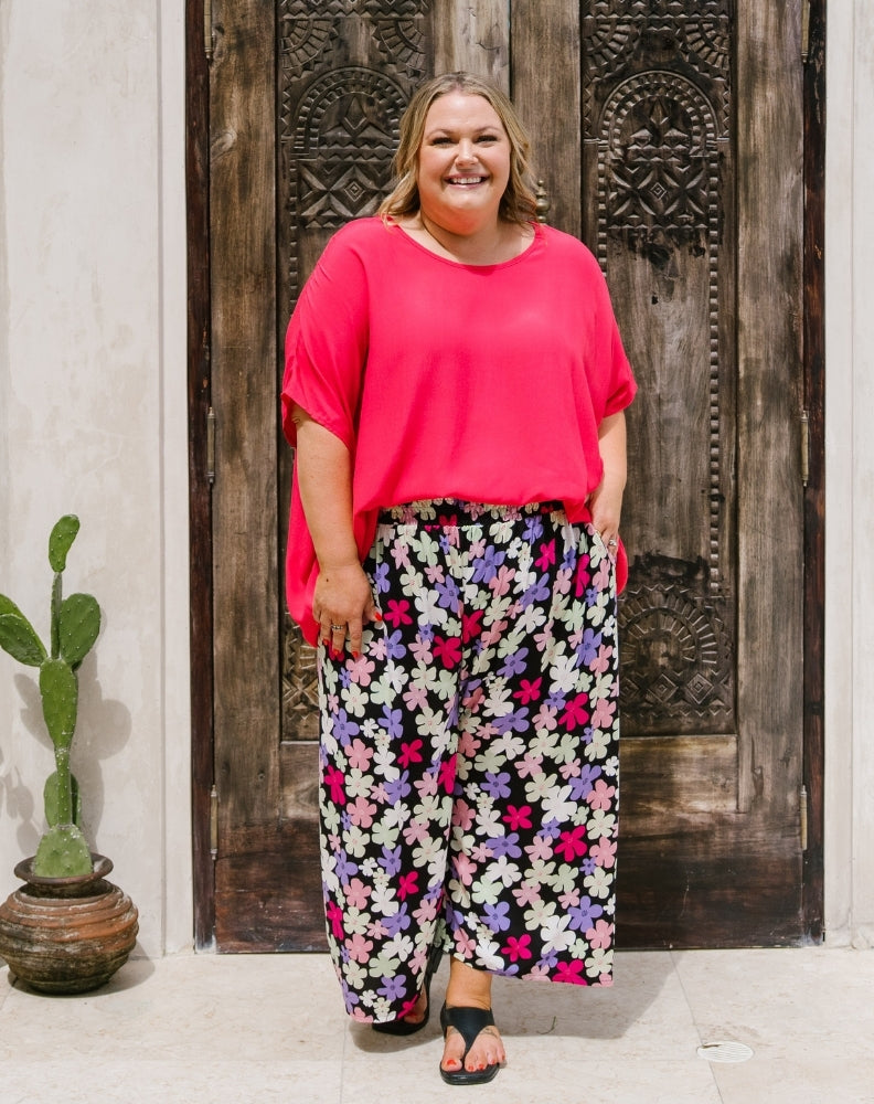 A woman stands in front of a carved dark wooden door wearing a bright pink short-sleeve top and black wide-leg pants with a colorful floral print. A small potted cactus sits on the left.